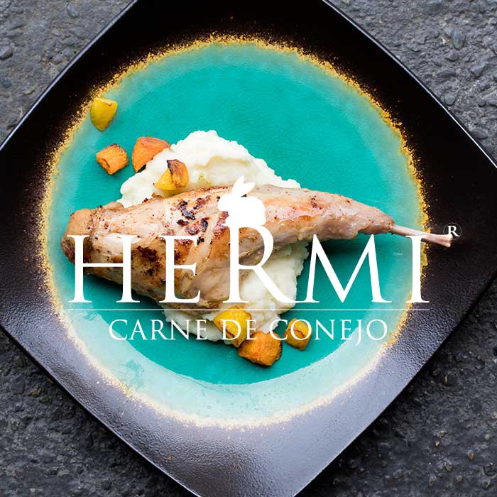 The Grupo Hermi logo superimpose over a cooked leg of Spanish rabbit with roasted mushrooms and mashed potatoes on a square black plate with a gold-rimmed turquoise circle in the middle. 
